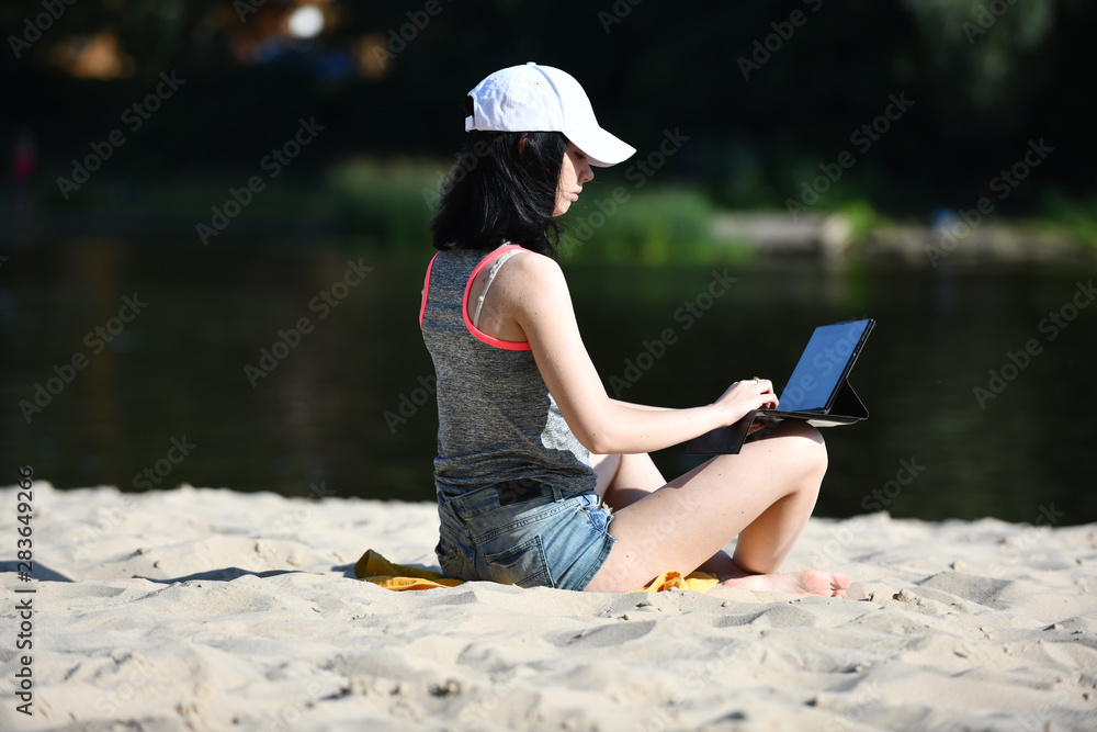 young woman sitting in jeans shorts and working with tablet on the beach