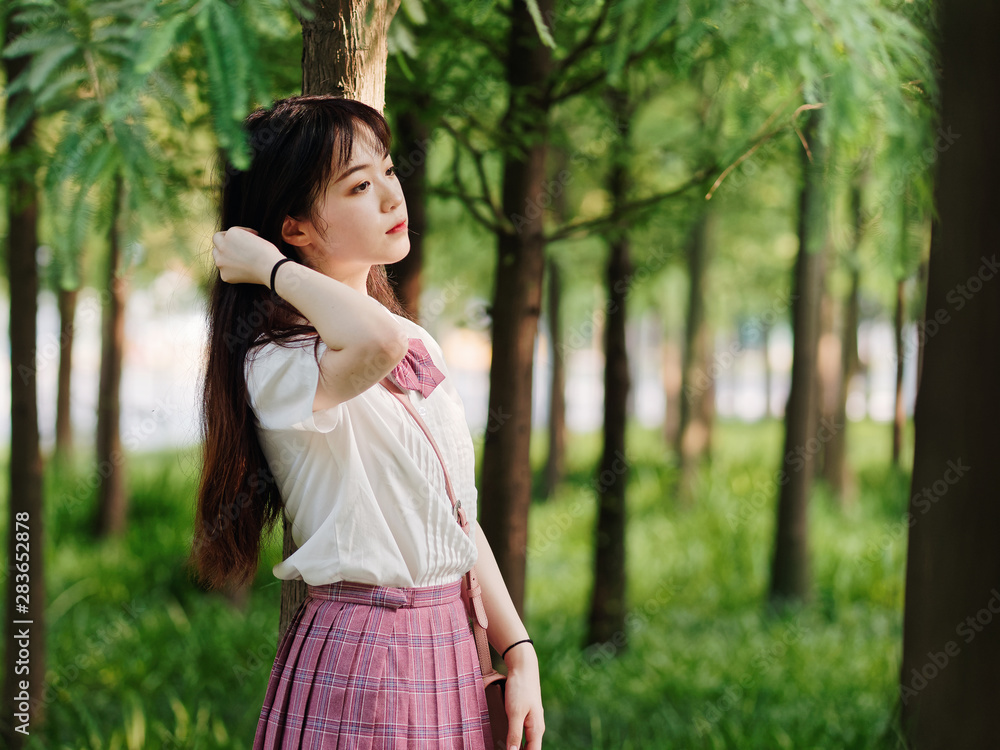 Portrait of beautiful Chinese girl in white shirt and pink skirt looking away with hand on hair in sunny summer forest, breathing the fresh air and enjoy nature. Beauty, lifestyle and summer Concept. Portrait of beautiful Chinese girl in white shirt and pink skirt looking away with hand on hair in sunny summer forest, breathing the fresh air and enjoy nature. Beauty, lifestyle and summer Concept.