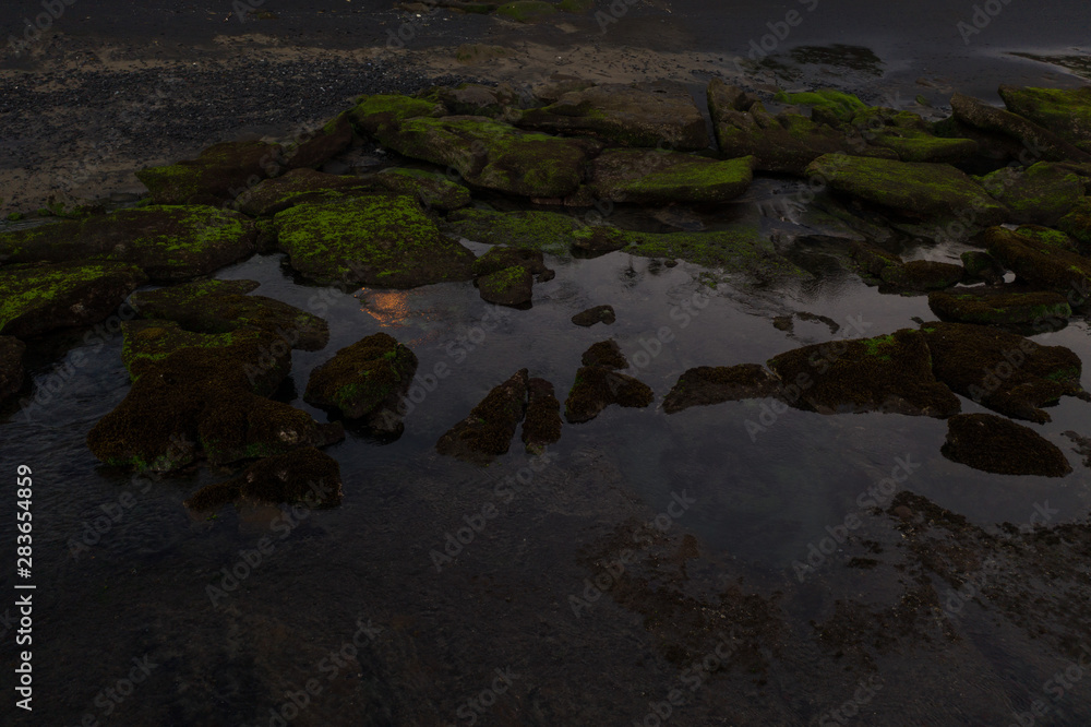 Aerial view of beautiful rocks covered by green moss in the sea near black sand beach during sunset Aerial view of beautiful rocks covered by green moss in the sea near black sand beach during sunset