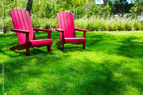 Traditional curveback sunset red plastic outdoor patio adirondack chairs with contoured backs and seats on green grass of outdoor lawn. Design, concept, idea