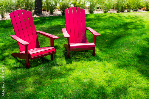 Traditional curveback sunset red plastic outdoor patio adirondack chairs with contoured backs and seats on green grass of outdoor lawn. Design, concept, idea © MichaelVi