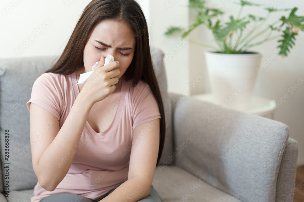 Asian Cute of girl having flu season and sneeze using paper tissues sitting on sofa at home, Health and illness concepts Asian Cute of girl having flu season and sneeze using paper tissues sitting on sofa at home, Health and illness concepts