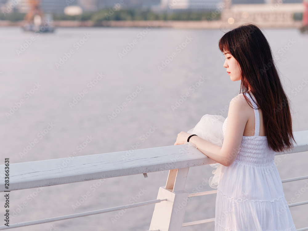 Portrait of beautiful Chinese girl in white bride dress standing at river bank and looking away in summer dusk, rear view, Beauty, lifestyle and summer Concept.