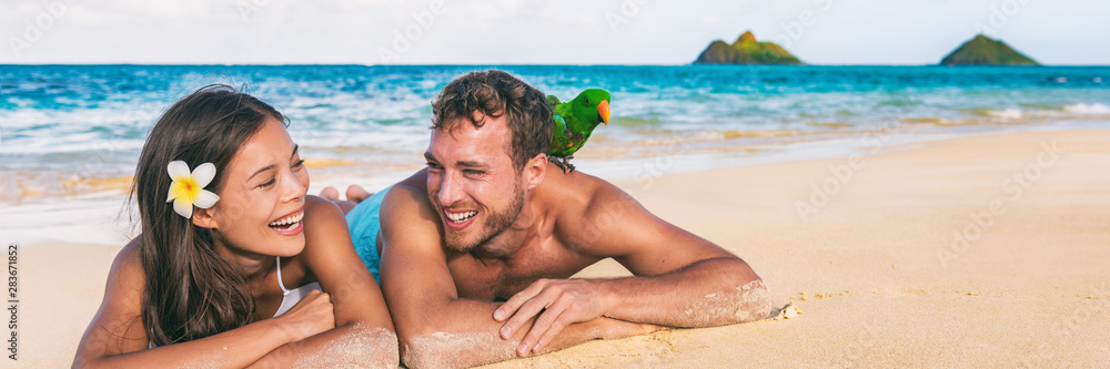 Fototapeta premium Beach people Hawaii honeymoon young couple laughing tourists with bird pet on shoulder -tourist attraction panoramic banner background, Lanikai, Oahu island, USA travel.