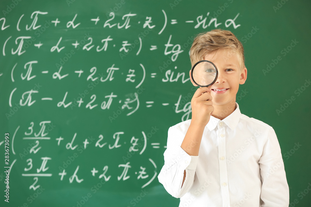 Little boy with magnifying glass near school blackboard in classroom