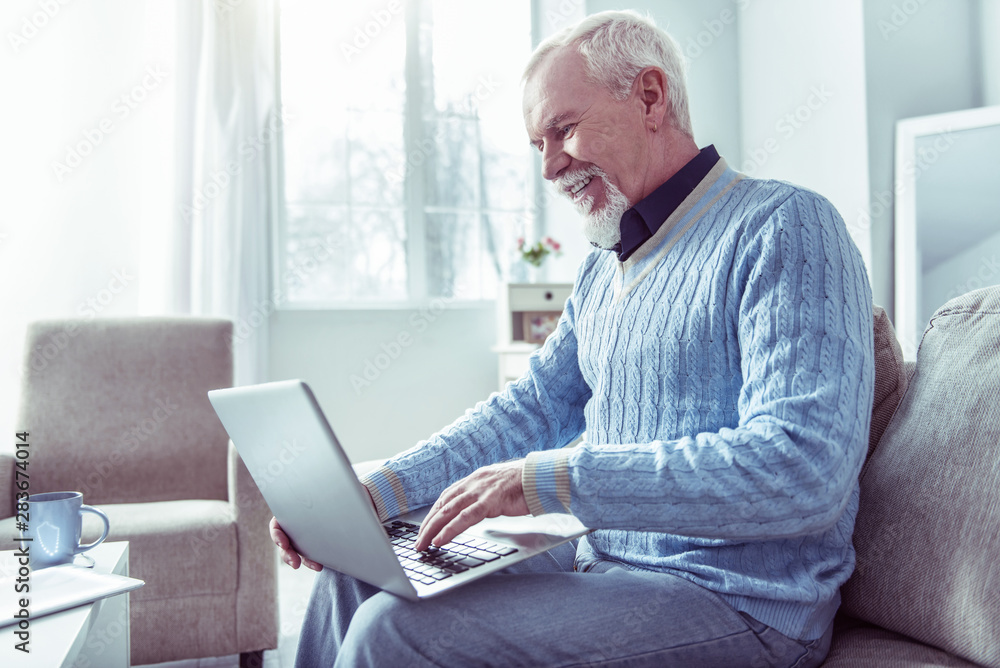 Beaming bearded retired man sitting on grey sofa having video chat Beaming bearded retired man sitting on grey sofa having video chat