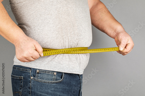 A man measures his fat belly with a measuring tape. on a gray background.