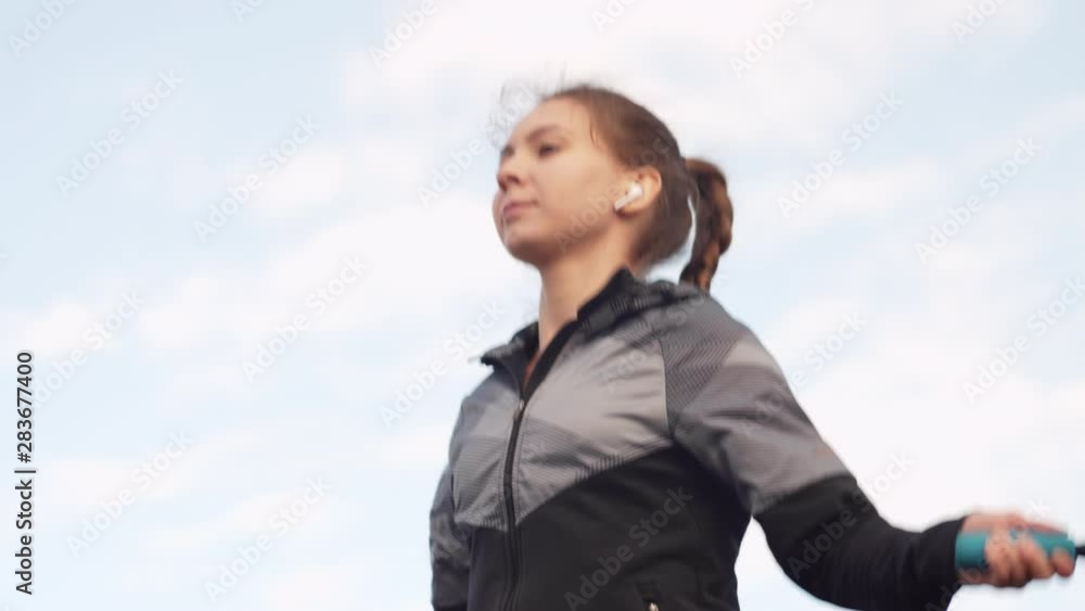 Tilt down of young Caucasian woman wearing sportswear listening to music using wireless headphones while skipping with jump rope on wooden embankment outside Tilt down of young Caucasian woman wearing sportswear listening to music using wireless headphones while skipping with jump rope on wooden embankment outside