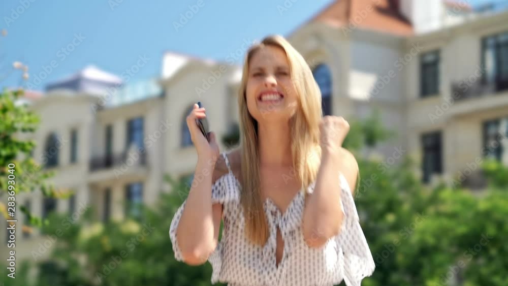 Happy blondy woman in dress using smartphone and rejoices while standing at park Happy blondy woman in dress using smartphone and rejoices while standing at park