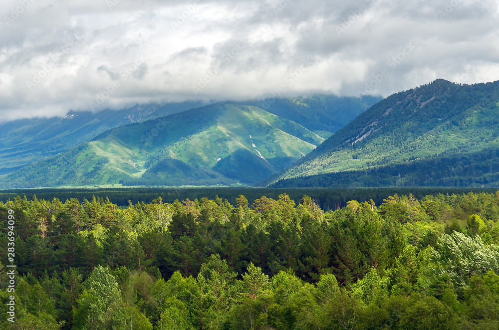 Sunbeams Shine through Heavy Clouds Flowing Low over the Forested Mountain Ridge and a Highland Valley on a Summer Afternoon. Altai Mountains, Kazakhstan. Sunbeams Shine through Heavy Clouds Flowing Low over the Forested Mountain Ridge and a Highland Valley on a Summer Afternoon. Altai Mountains, Kazakhstan.