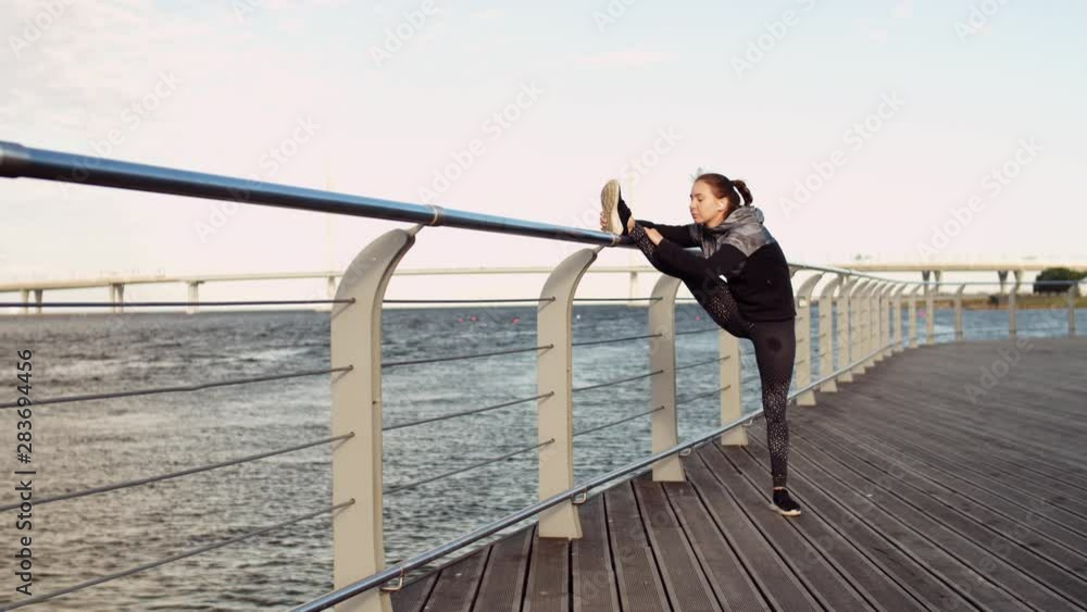 Wide shot of slim Caucasian female athlete standing on wooden embankment with her right leg on railing and doing stretching exercise Wide shot of slim Caucasian female athlete standing on wooden embankment with her right leg on railing and doing stretching exercise