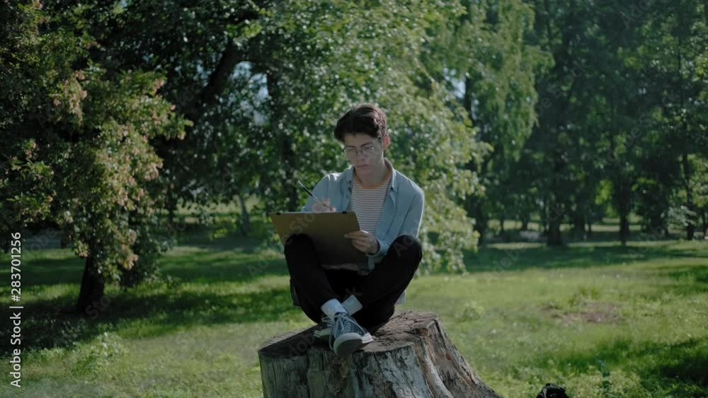 A young creative guy in round glasses and a denim shirt sits in a city park on a stump and draws into an album. Plein air: an artist alone paints nature in a summer park. A young creative guy in round glasses and a denim shirt sits in a city park on a stump and draws into an album. Plein air: an artist alone paints nature in a summer park.