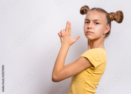 Portrait of funny teen girl doing gun gesture getting ready to shoot. Caucasian young teenager showing raised gun gesture on gray background. Child holding symbolic gun. Emotions and signs.