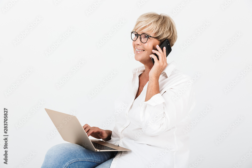 Image of mature adult woman wearing eyeglasses talking on smartphone while sitting with laptop computer