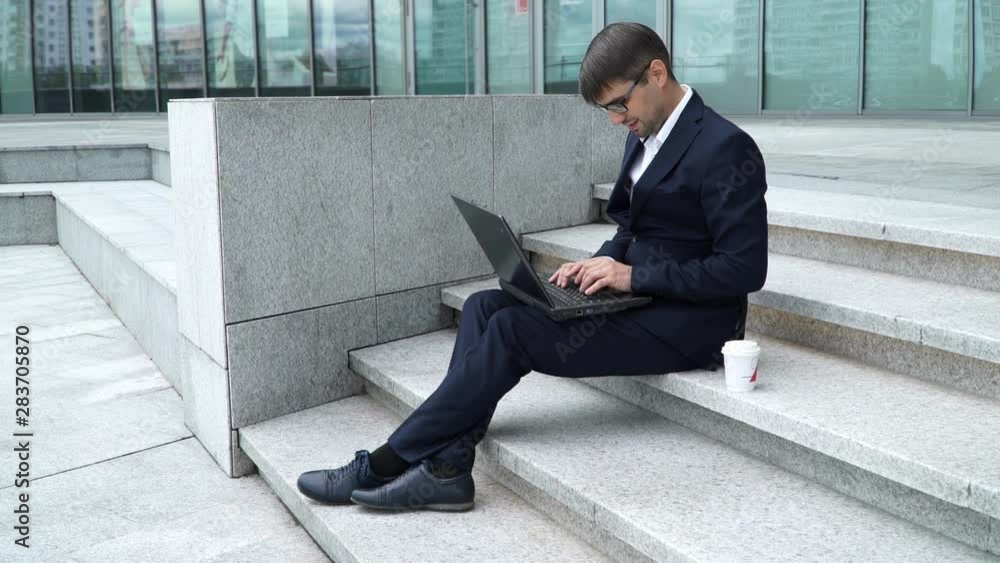 Portrait of young smiling businessman in glasses in suit sits on steps on background of business center and types on laptop, next is glass of coffee. Man works outdoors on computer. Portrait of young smiling businessman in glasses in suit sits on steps on background of business center and types on laptop, next is glass of coffee. Man works outdoors on computer.