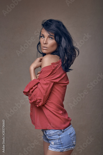 Beautiful sexy woman in skimpy denim shorts with her shirt slipped down provocatively off her shoulder looking at the camera with parted lips over a brown studio background