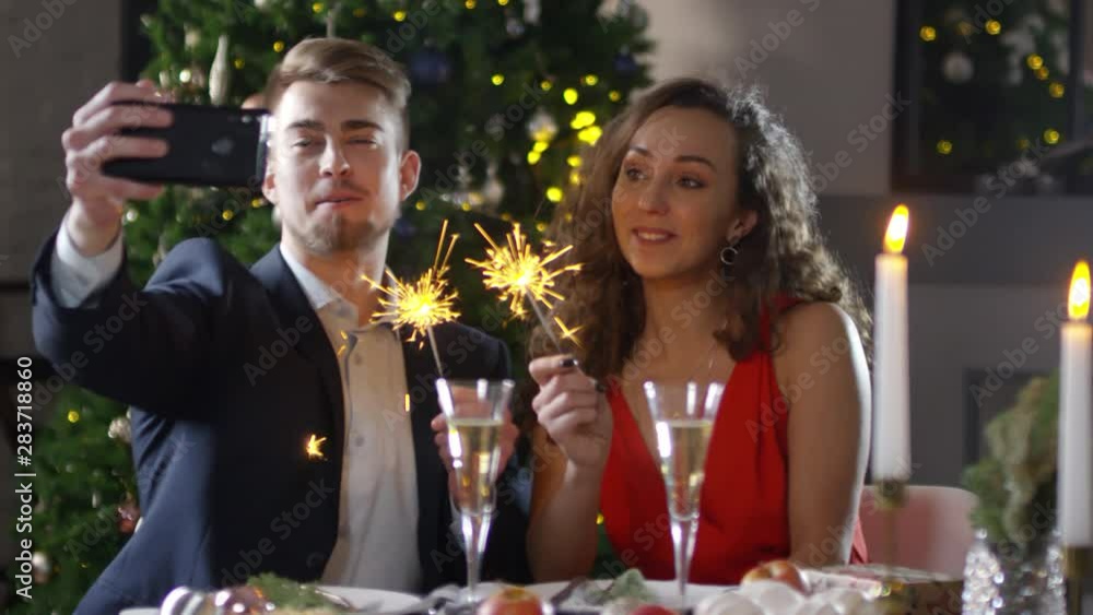Medium shot of lovely young Caucasian couple sitting together at festive dinner table in front of decorated Christmas tree with burning sparklers and taking selfies on smartphone, smiling and talking Medium shot of lovely young Caucasian couple sitting together at festive dinner table in front of decorated Christmas tree with burning sparklers and taking selfies on smartphone, smiling and talking