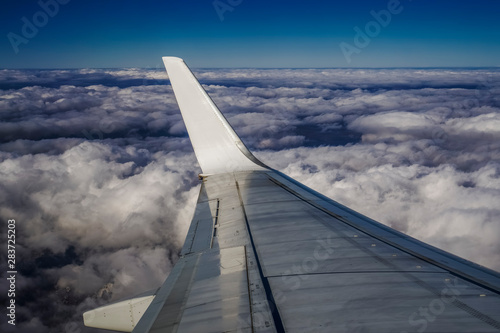 Window wing view of airplane flying on high altitude  above a bed of puffy clouds. Day view inside commercial plane window of a metal wing  without carrier logo.