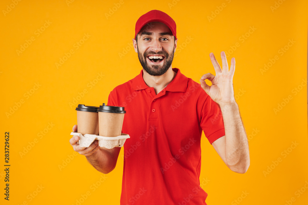 Portrait of unshaven delivery man in red uniform showing ok sign and holding takeaway coffee cups Portrait of unshaven delivery man in red uniform showing ok sign and holding takeaway coffee cups