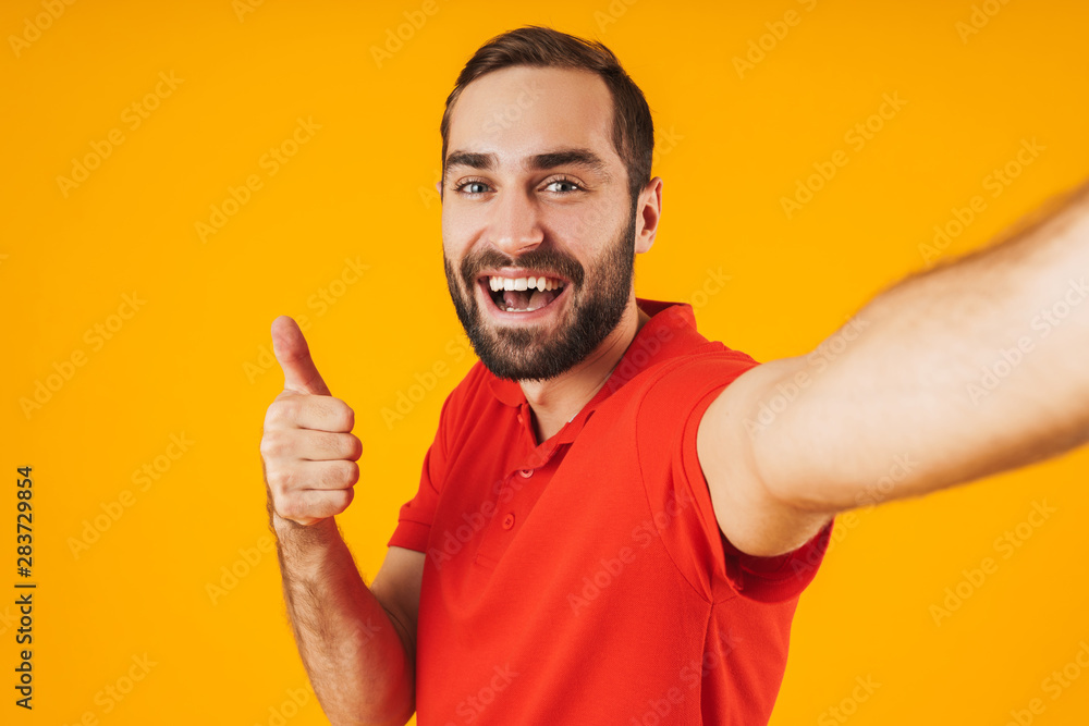 Portrait of delighted man in red t-shirt laughing and showing thumb up while taking selfie photo isolated over yellow background Portrait of delighted man in red t-shirt laughing and showing thumb up while taking selfie photo isolated over yellow background