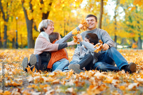 Happy family sitting on fallen leaves, playing and having fun in autumn city park. Children and parents together having a nice day. Bright sunlight and yellow leaves on trees, fall season.