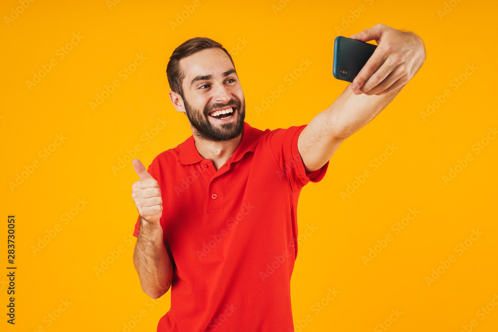 Portrait of attractive man in red t-shirt laughing and showing thumb up while taking selfie photo on cellphone Portrait of attractive man in red t-shirt laughing and showing thumb up while taking selfie photo on cellphone