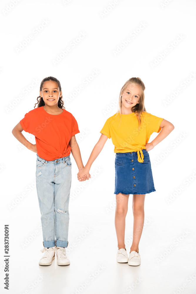 full length view of two cheerful multicultural friends holding hands while smiling at camera on white background full length view of two cheerful multicultural friends holding hands while smiling at camera on white background