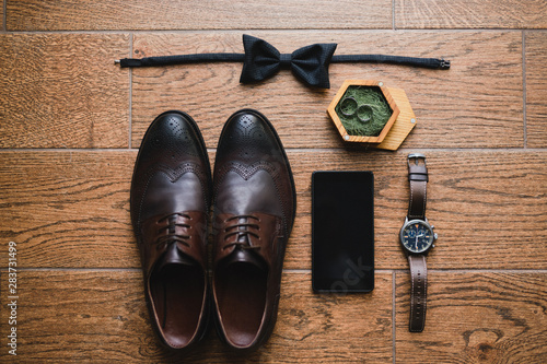 Wedding fashion for men. Top view of modern groom's outfit: bow tie, smart phone, wedding rings, watch, leather shoes and boutonniere on wooden background.