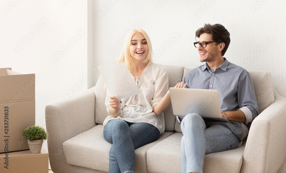Couple reading certificate of property sitting on sofa using laptop Couple reading certificate of property sitting on sofa using laptop