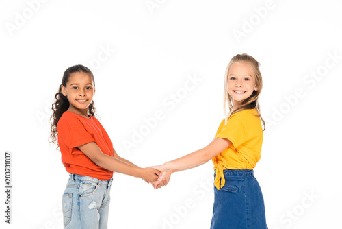 two cute multicultural friends holding hands and smiling at camera isolated on white