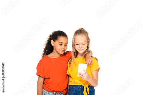 cheerful african american kid hugging smiling friend using smartphone isolated on white