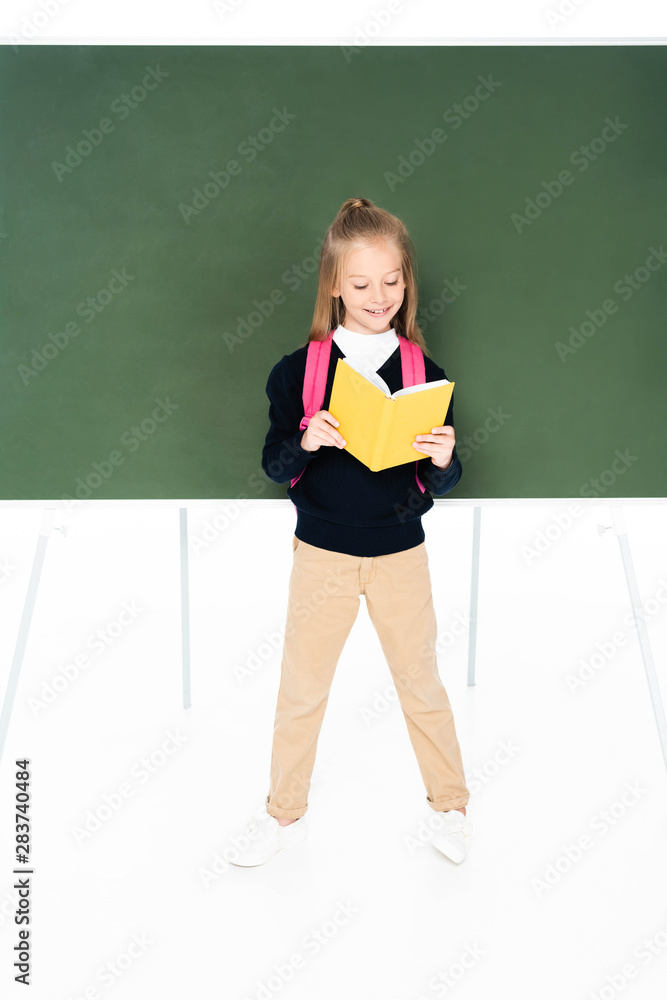 full length view of schoolgirl reading book while standing near green chalkboard on white background full length view of schoolgirl reading book while standing near green chalkboard on white background