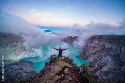 Man traveler standing on edge of crater with colorful sky at morning. Beautiful Ijen volcano with acid lake and sulfur gas going from crater, Indonesia photo