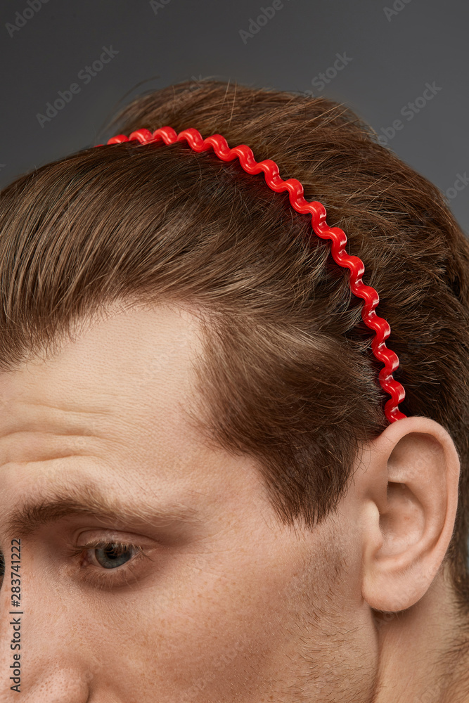 Naklejka premium Extreme close-up shot of a young chestnut European man with a fire red hairband in the form of a wavy wire in his hair. The fashion model with an unshaved cheek-bone is looking away. 