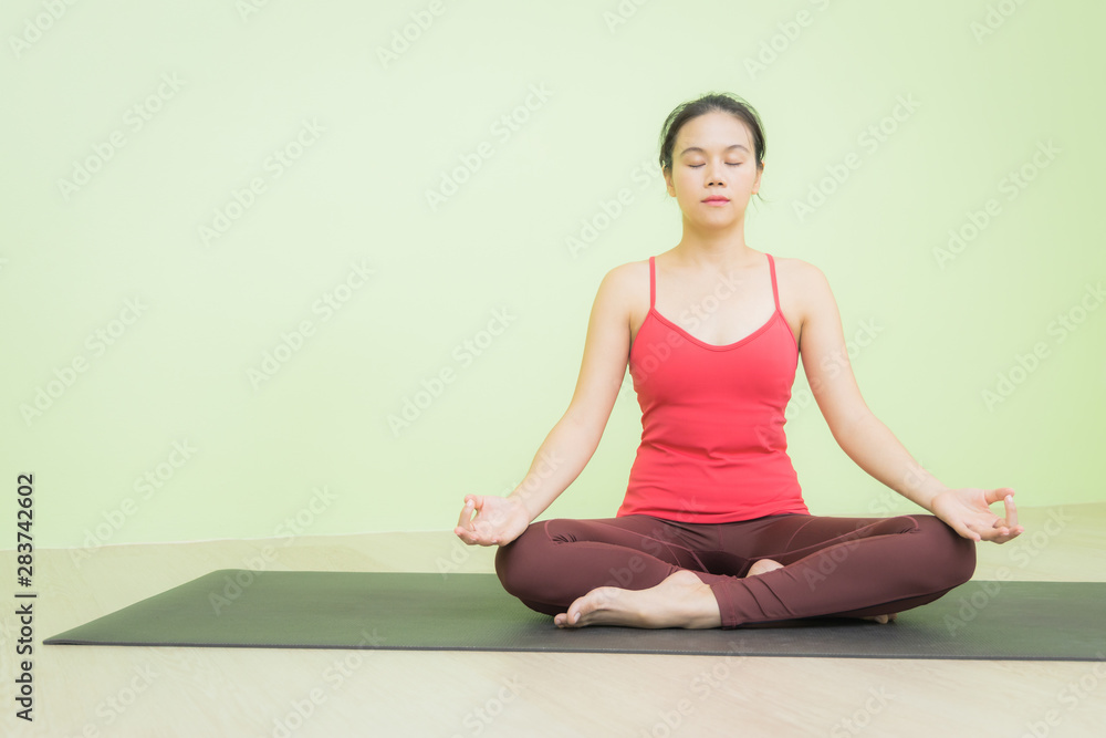 Happy and peacefully young Asian woman in bright red cloth sitting on black yoga mat practicing meditation in Lotus pose yoga.