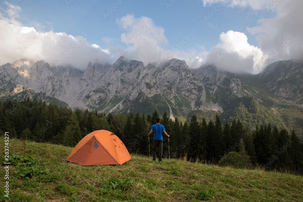 Male hiker standing near his trekking tent on a green and beautiful mountain  landscape. Adventure travel lifestyle concept