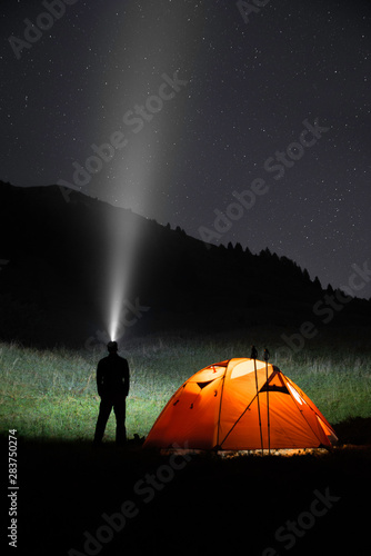 Silhouette of a hiker looking up at the sky full of stars with a headlight in a dark night. Mountain landscape with an illuminated camping tent