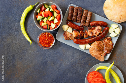 Traditional Serbian and Balkan grilled meat called mesano meso. Balkan barbeque (rostilj) served with Serbian salad, hot peppers, bread, tomato, onions, and paprika powder. Dark background. Top view photo