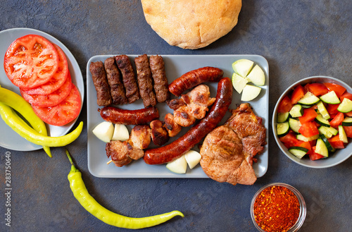 Traditional Serbian and Balkan grilled meat called mesano meso. Balkan barbeque (rostilj) served with Serbian salad, hot peppers, bread, tomato, onions, and paprika powder. Dark background. Top view photo