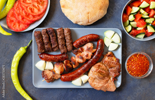 Traditional Serbian and Balkan grilled meat called mesano meso. Balkan barbeque (rostilj) served with Serbian salad, hot peppers, bread, tomato, onions, and paprika powder. Dark background. Top view photo
