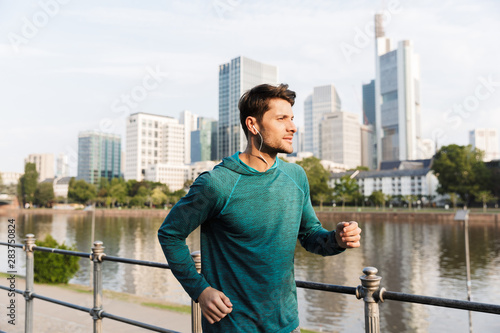 Photo of calm serious man listening music with earphones while running near city promenade