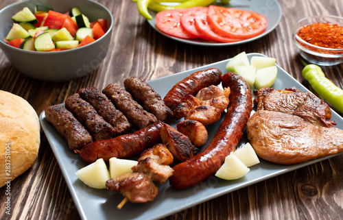Traditional Serbian and Balkan grilled meat called mesano meso. Balkan barbeque (rostilj) served with Serbian salad, hot peppers, bread, tomato, onions, and paprika powder. Wooden background. Close-up