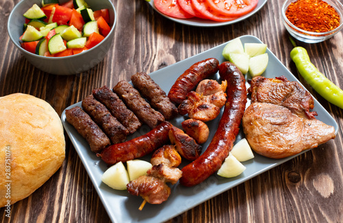 Traditional Serbian and Balkan grilled meat called mesano meso. Balkan barbeque (rostilj) served with Serbian salad, hot peppers, bread, tomato, onions, and paprika powder. Wooden background. Close-up photo