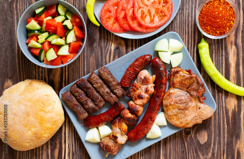 Traditional Serbian and Balkan grilled meat called mesano meso. Balkan barbeque (rostilj) served with Serbian salad, hot peppers, bread, tomato, onions, and paprika powder. Wooden background. Top view photo