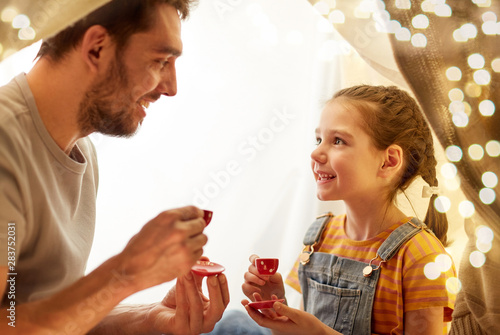 family, hygge and people concept - happy father and little daughter playing tea party in kids tent at night at home