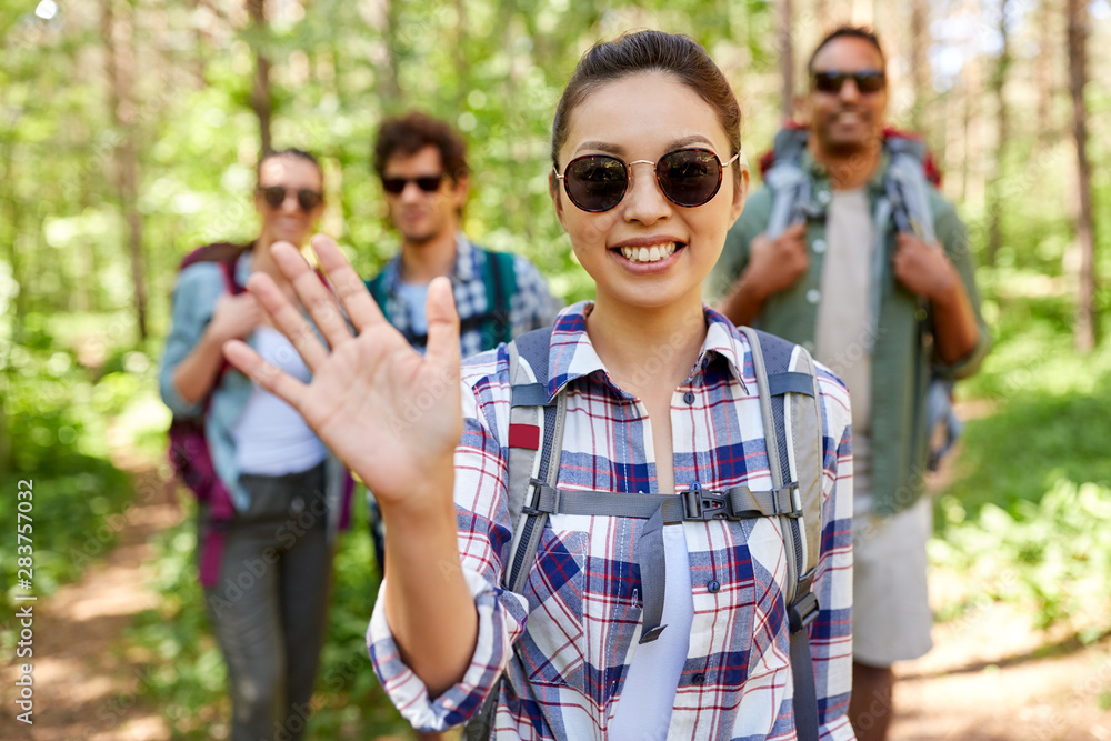 travel, tourism, hike and people concept - group of friends with backpacks and asian woman waving hand in forest
