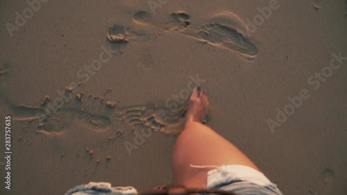 Top view close-up of young woman legs standing on sea shore at sunset. Foamy sea waves gentrly wash sand and feet, warm soft evening sun reflects on wet surface. First person view slow motion footage.