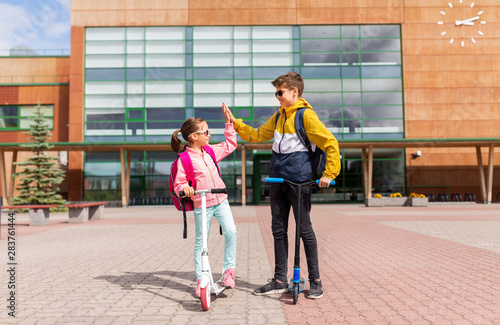 education  childhood and people concept - happy school children with backpacks riding scooters and making high five outdoors