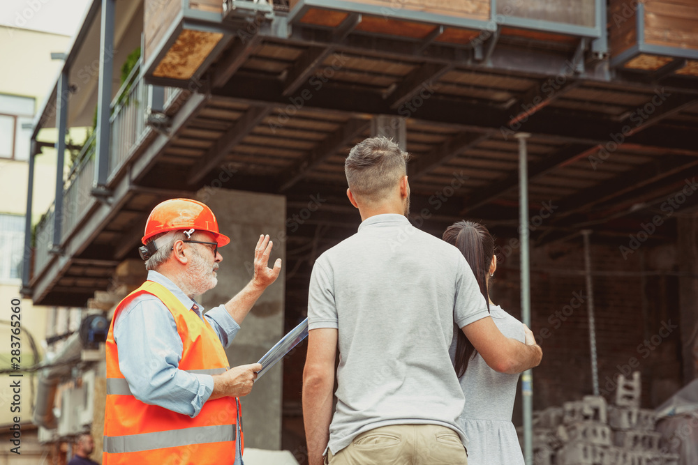 Foreman or achitect engineer shows future house, office or store design plans to a young couple. Meeting at the construction site to talk about facade appearance, interior decoration, home layout. Foreman or achitect engineer shows future house, office or store design plans to a young couple. Meeting at the construction site to talk about facade appearance, interior decoration, home layout.