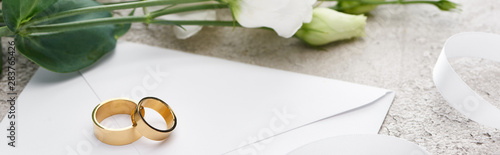 panoramic shot of white envelope with golden rings near white flowers on textured surface